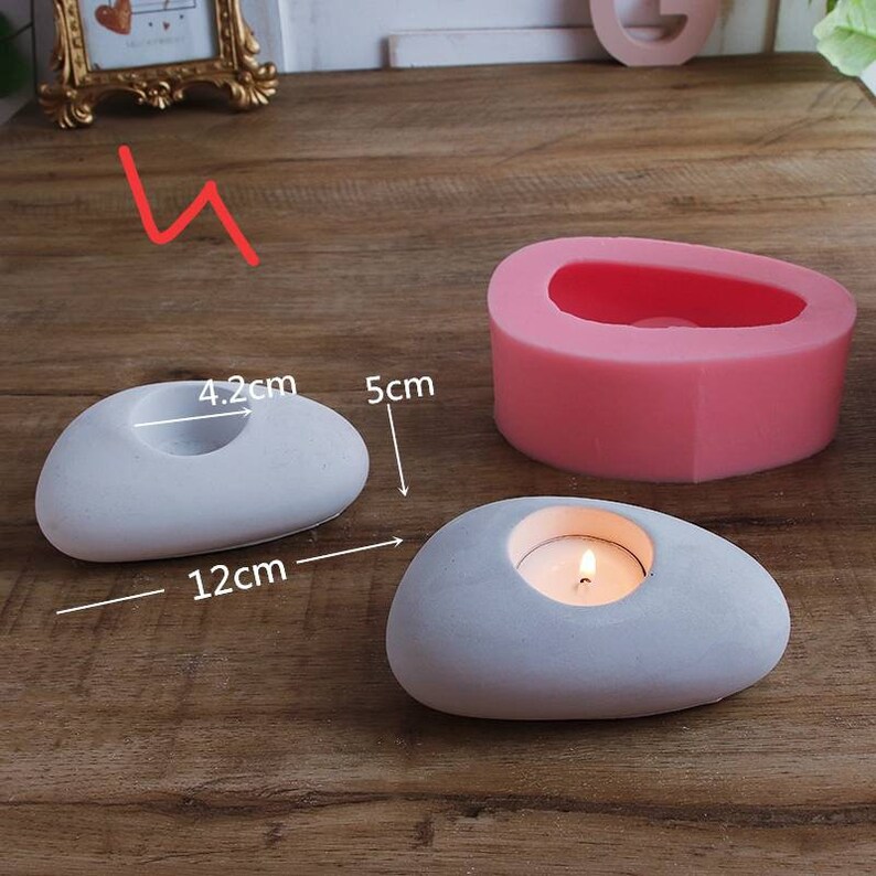May include: Two gray stone-shaped candle holders with a round opening for a tea light candle. The larger holder measures 12 inches long and 5 inches tall. The smaller holder measures 4.2 inches wide. A pink silicone mold for making the candle holders is also shown.
