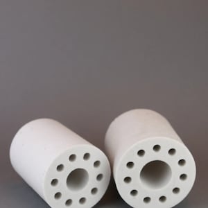 Op de afbeelding: Twee witte keramische cilinders met meerdere kleine gaten rond de omtrek. De cilinders zijn hol en hebben een groter gat in het midden.