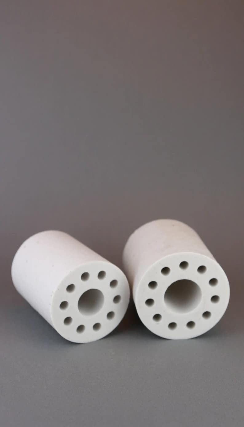 Op de afbeelding: Twee witte keramische cilinders met meerdere kleine gaten rond de omtrek. De cilinders zijn hol en hebben een groter gat in het midden.