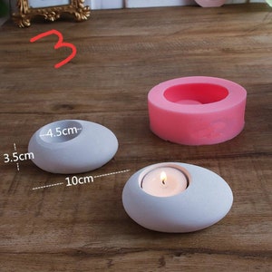 May include: Two white stone candle holders in the shape of a pebble with a 4.5 cm diameter hole for a tea light candle. The larger holder measures 10 cm in length and 3.5 cm in height. A pink silicone mold for the candle holder is also shown.
