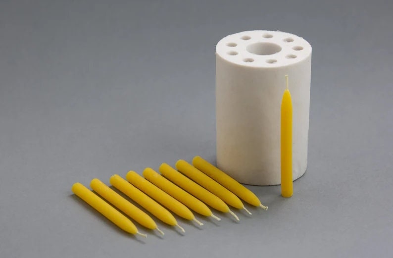 Op de afbeelding: Een witte siliconen mal met 10 ronde gaten voor het maken van kaarsen. Tien gele bijenwaskaarsen zijn in een rij naast de mal gerangschikt.