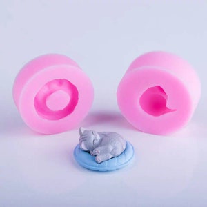 Op de afbeelding: Twee roze siliconen mallen om een ​​slapende kattenfiguur te maken. De mallen zijn rond en hebben een hol midden. Een klein grijs kattenfiguur is te zien terwijl het slaapt op een blauw kussen.