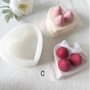 May include: A white heart-shaped silicone mold with a smaller heart-shaped cavity inside. Next to it are two heart-shaped cakes decorated with pink and red strawberries.
