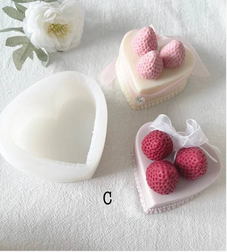 May include: A white heart-shaped silicone mold with a smaller heart-shaped cavity inside. Next to it are two heart-shaped cakes decorated with pink and red strawberries.