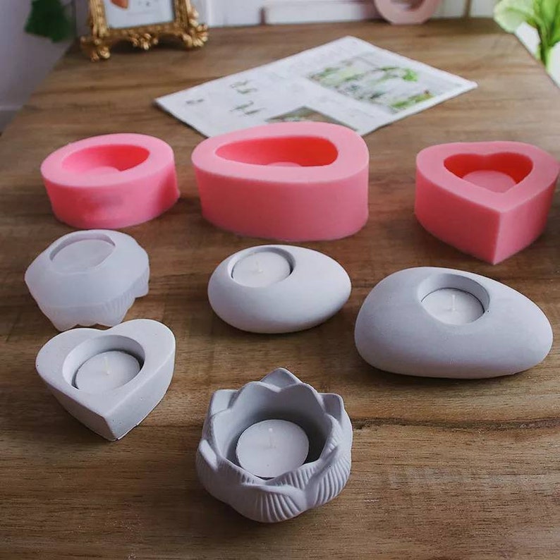 May include: A set of eight silicone molds for making concrete candle holders. The molds are in the shapes of a heart, a flower, a pebble, and a rectangle. The molds are pink and gray.