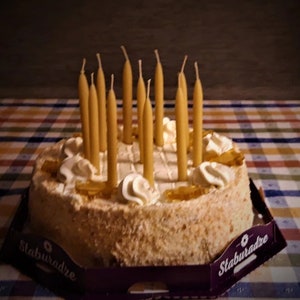 Op de afbeelding: Een ronde cake met witte frosting en verschillende toppings, waaronder karamel en slagroom. De cake is versierd met 15 gele kaarsen.