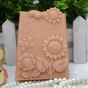 May include: A light brown rectangular soap mold with a raised design of sunflowers, butterflies, and leaves. The mold has the text "Hello, Sunshine!" embossed on the front.