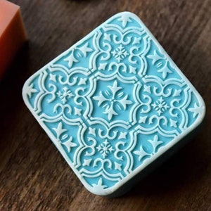 K&ouml;nnte beinhalten: Eine quadratische, hellblaue Seife mit einem wei&szlig;en, filigranen, geometrischen Muster, das auf der Oberseite eingepr&auml;gt ist.