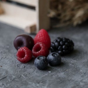 May include: A close-up of a selection of fresh berries, including red raspberries, a red strawberry, a dark purple cherry, and two dark blue blueberries. The berries are arranged on a grey textured surface.