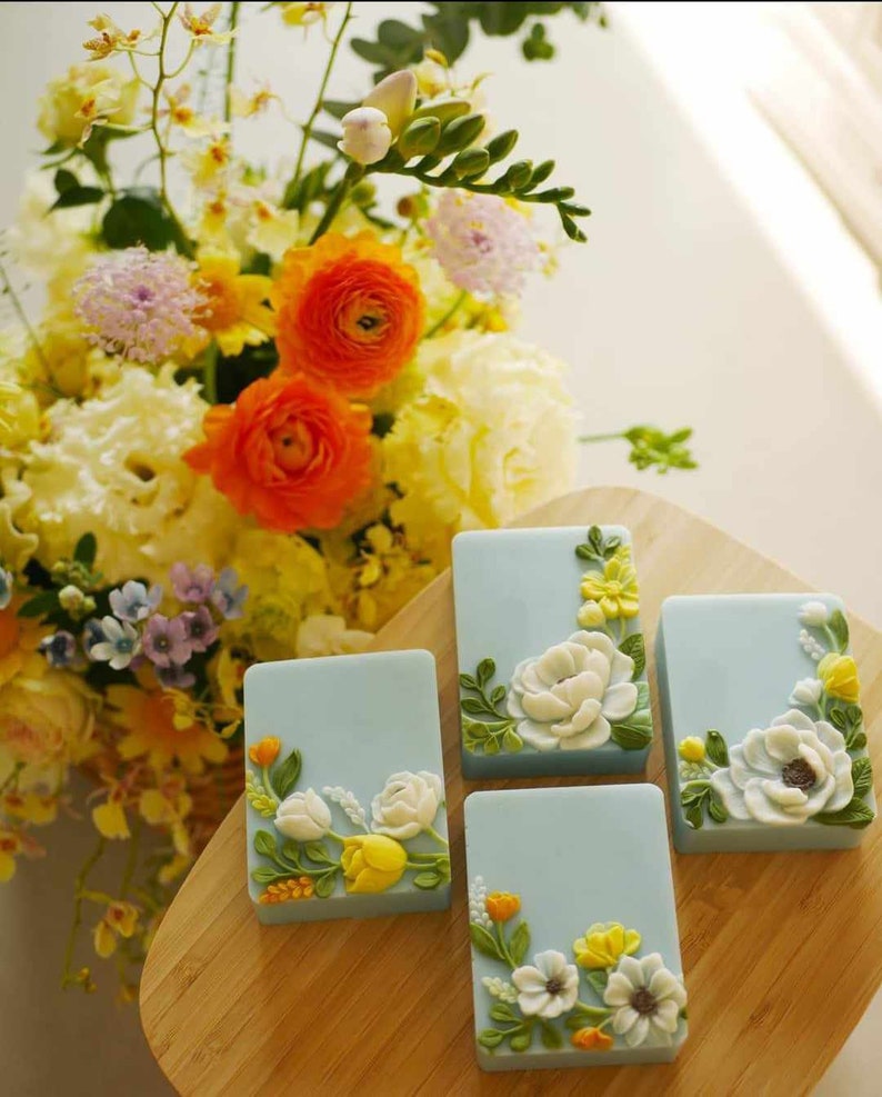 May include: Four square blue soap bars with floral designs. Each bar features a white flower with yellow and green accents.