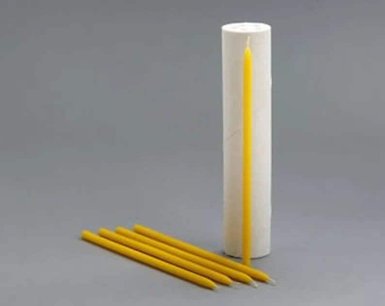 Puede incluir: Un juego de seis velas de cera de abeja amarilla, incluida una vela larga y delgada en un tubo de papel blanco. Las velas est&aacute;n dispuestas en forma de abanico sobre una superficie gris.