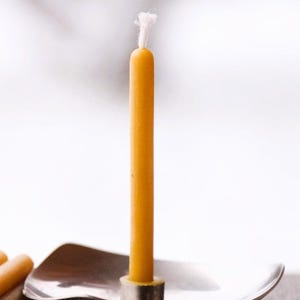 Op de afbeelding: Een enkele gele bijenwaskaars met een witte lont staat in een zilveren kandelaar. De kandelaar heeft een gebogen, ondiepe schotelvorm met een ronde basis.
