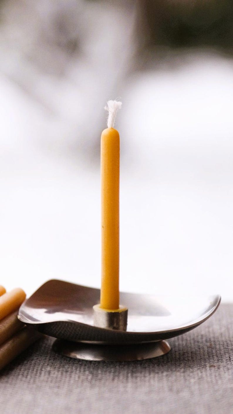 Op de afbeelding: Een enkele gele bijenwaskaars met een witte lont staat in een zilveren kandelaar. De kandelaar heeft een gebogen, ondiepe schotelvorm met een ronde basis.