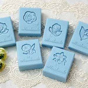 Könnte beinhalten: Sechs hellblaue, rechteckige handgemachte Seifen mit geprägten Blumendesigns, darunter Rosen und Lilien. Jede Seife trägt das Wort "Handmade" in Schreibschrift. Die Seifen sind auf einer weißen Spitzendecke angeordnet.