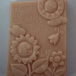 May include: A light brown rectangular silicone mold with a raised design of a sun, flowers, leaves, a butterfly, and a bird. The mold has the text "Have a Sunny Day" inscribed on it.