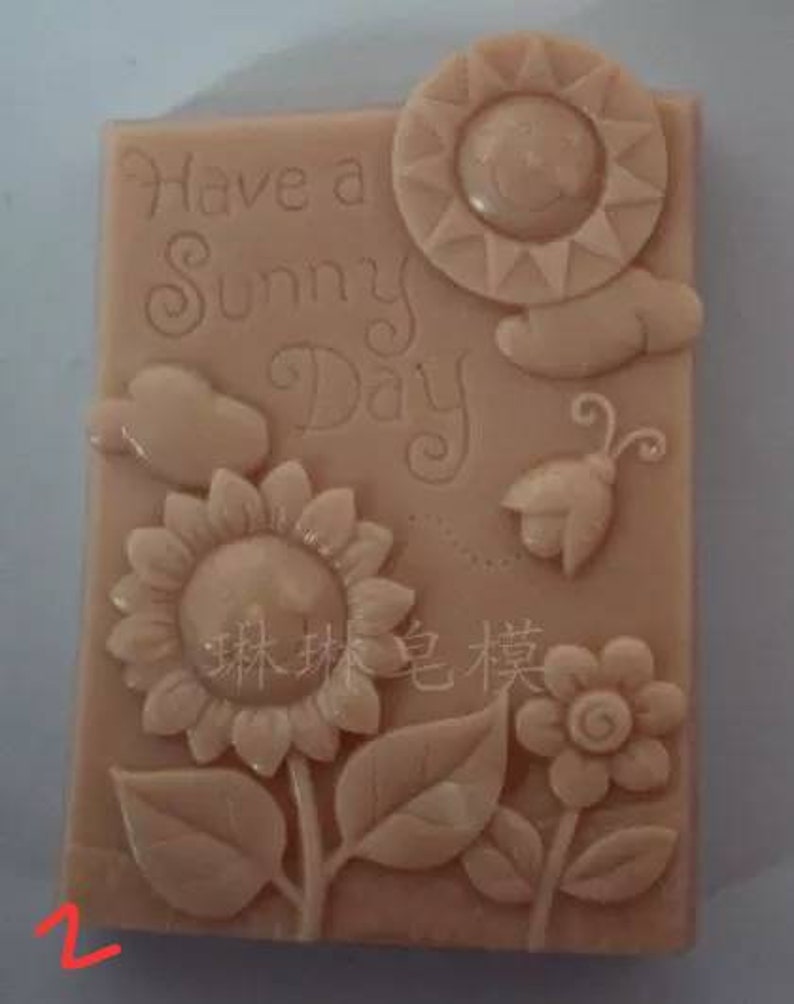 May include: A light brown rectangular silicone mold with a raised design of a sun, flowers, leaves, a butterfly, and a bird. The mold has the text "Have a Sunny Day" inscribed on it.