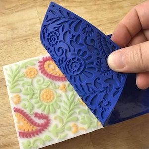 Könnte beinhalten: Ein blauer Silikon-Kuchen-Dekor-Stempel mit einem floralen Design. Der Stempel wird verwendet, um ein geprägtes Design auf einer weißen Fondant-Oberfläche zu erstellen.