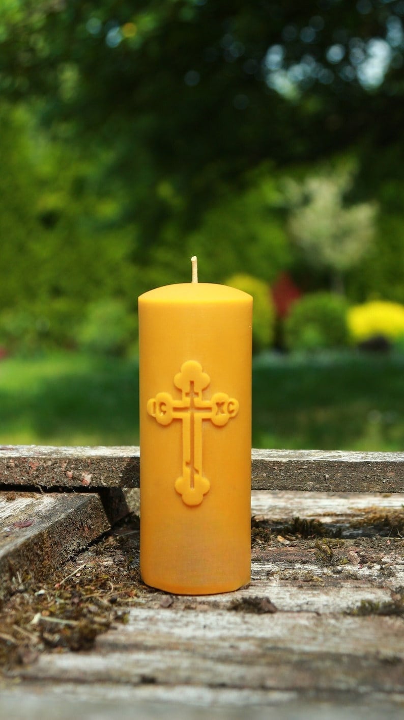 May include: A yellow beeswax candle with a cross design and the inscription "IC XC" on the front. The candle is sitting on a wooden surface.