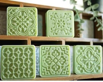 May include: Six square-shaped green soap bars with different geometric patterns. The soap bars are arranged in two rows of three on a wooden shelf.