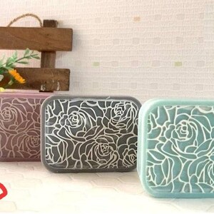 K&ouml;nnte beinhalten: Drei rechteckige Plastikbeh&auml;lter mit Blumendesigns. Die Beh&auml;lter sind in den Farben Rosa, Grau und Hellblau. Das Blumendesign ist ein sich wiederholendes Muster aus Rosen.