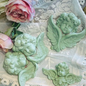 May include: Four decorative cherub figurines with wings, made of a light green material, are arranged on a white lace background. The cherubs have delicate features and are adorned with floral details.