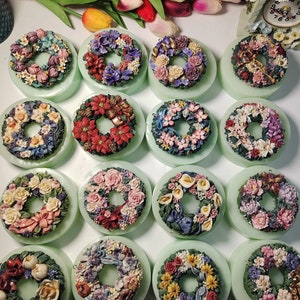 May include: A collection of 15 handmade wreaths with various floral designs, each displayed on a green base. The wreaths feature a variety of colours, including red, pink, yellow, blue, and green.