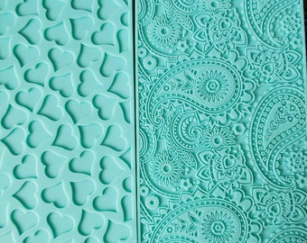 Könnte beinhalten: Zwei türkisfarbene Silikon-Backmatten mit unterschiedlichen Mustern. Eine Matte hat ein sich wiederholendes Herz-Design, die andere ein Paisley-Muster.