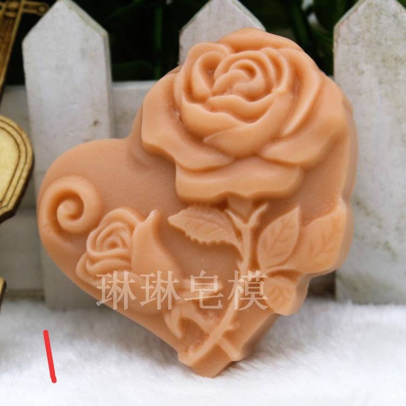 May include: A heart-shaped silicone mold with a rose design. The mold is light brown and features a large rose with leaves and a smaller rose with a swirl design.