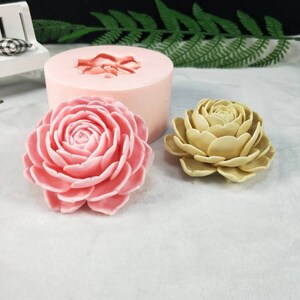 May include: Two silicone molds for making flower-shaped candles. One mold is pink and the other is beige. The molds are on a white surface.