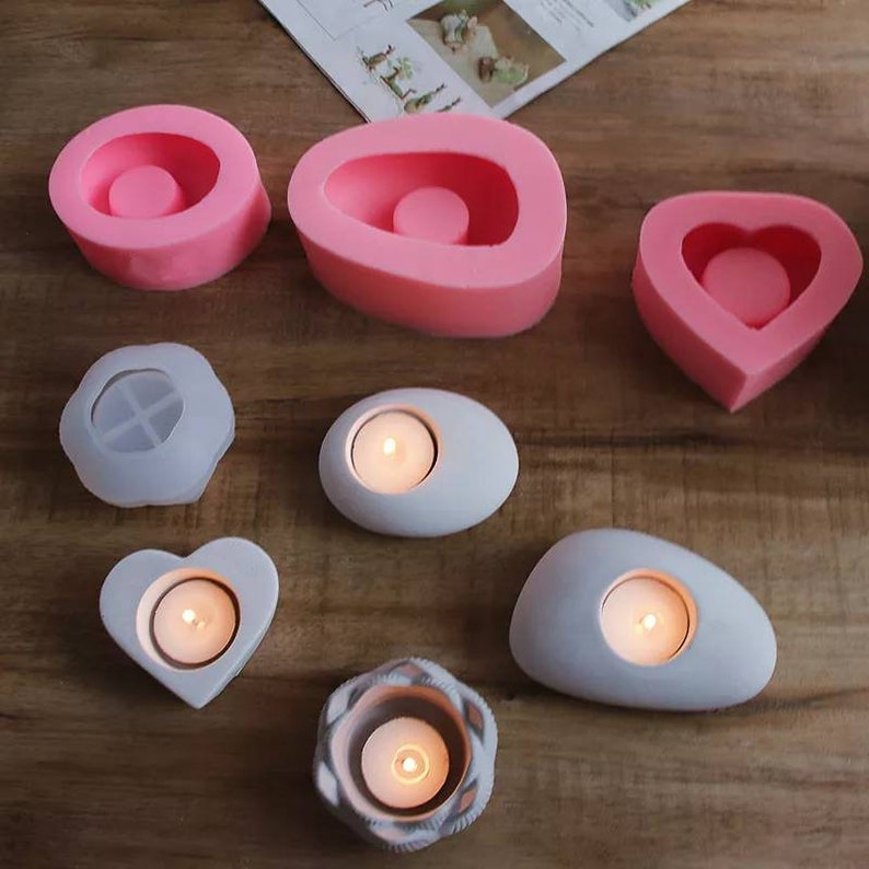 May include: Assortment of pink silicone candle molds and white concrete candle holders in various shapes, including heart, oval, and round. The concrete candle holders have lit tea lights inside.