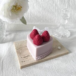 May include: A heart-shaped cake decorated with three red strawberries and a white ribbon. The cake is on a light brown square base.