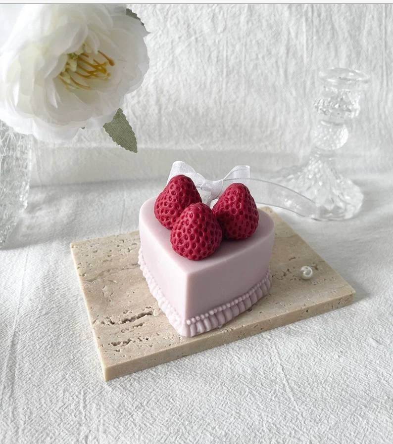 May include: A heart-shaped cake decorated with three red strawberries and a white ribbon. The cake is on a light brown square base.