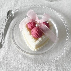 May include: A white heart-shaped cake with pink frosting and three pink strawberries on top. The cake is decorated with white frosting and a pink ribbon.