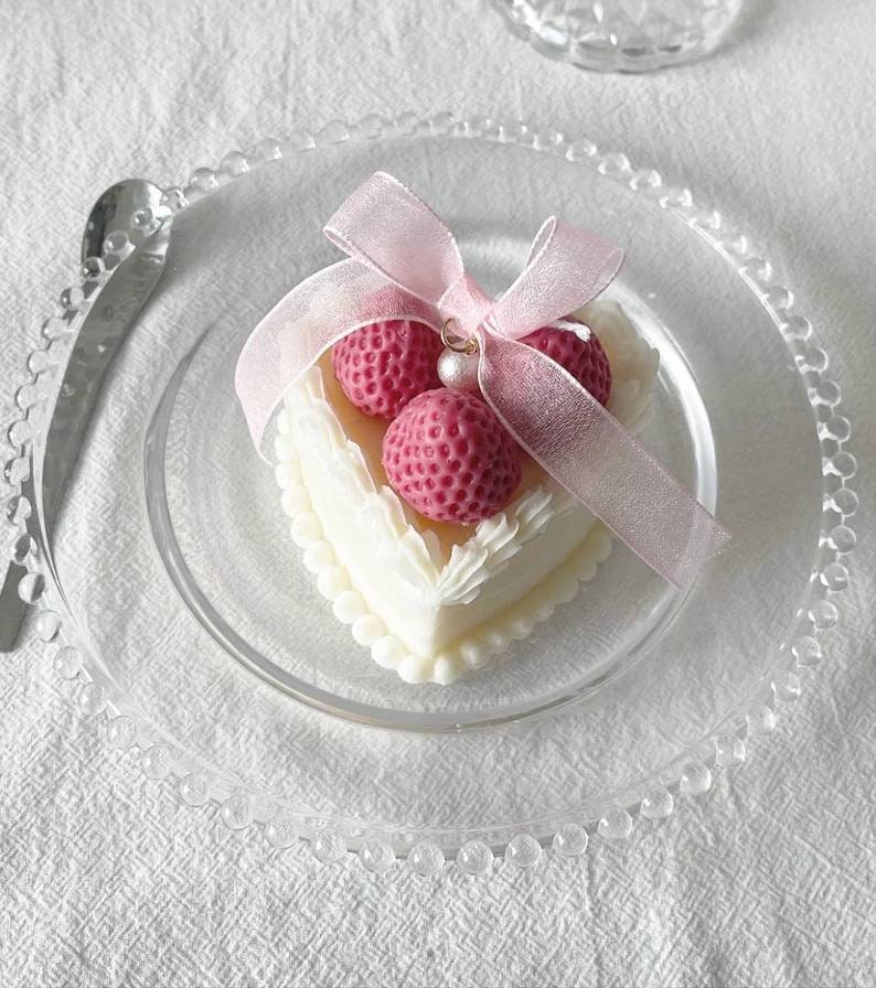 May include: A white heart-shaped cake with pink frosting and three pink strawberries on top. The cake is decorated with white frosting and a pink ribbon.