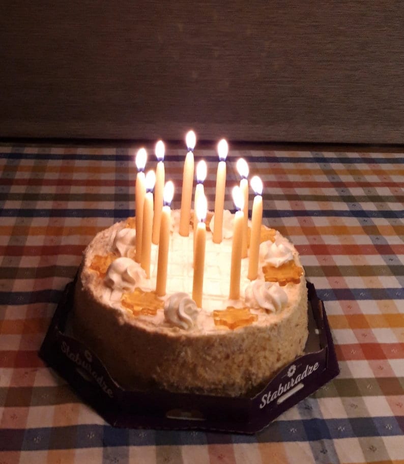 Op de afbeelding: Een ronde cake met witte frosting en verschillende toppings, waaronder karamel en noten. De cake is versierd met 18 brandende kaarsen.
