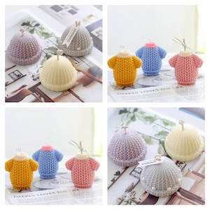 Könnte beinhalten: Satz mit sechs handgefertigten Kerzen in Form von gestrickten Mützen und Pullovern. Die Kerzen sind in verschiedenen Farben, darunter Gelb, Rosa, Blau und Grau. Die Kerzen sind mit kleinen weißen Perlen verziert und haben ein "Handmade"-Etikett.