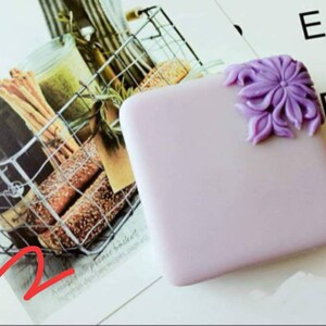 K&ouml;nnte beinhalten: Ein kleines, quadratisches, lavendelfarbenes Etui mit einem dekorativen violetten Blumendesign an der Ecke. Das Etui liegt auf einer wei&szlig;en Oberfl&auml;che.