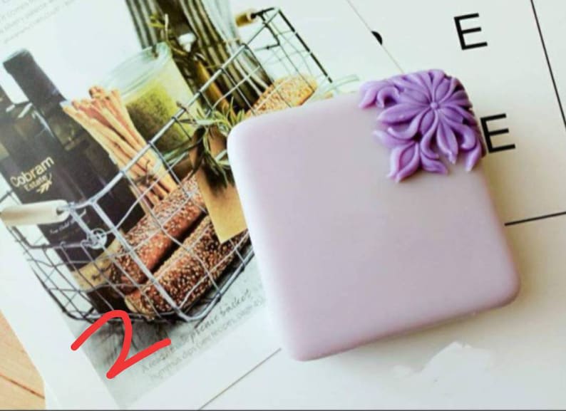 K&ouml;nnte beinhalten: Ein kleines, quadratisches, lavendelfarbenes Etui mit einem dekorativen violetten Blumendesign an der Ecke. Das Etui liegt auf einer wei&szlig;en Oberfl&auml;che.