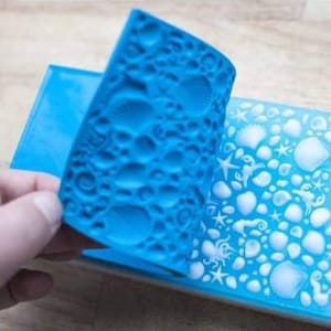 Pode incluir: Um molde de silicone azul com um design em relevo de conchas e outros elementos com tema oceânico. O molde é flexível e pode ser usado para criar sabão, resina ou outros artesanatos.