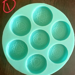 Puede incluir: Un molde de silicona turquesa con siete impresiones circulares. Cada impresión presenta un símbolo de chakra diferente, un diseño geométrico que representa un centro de energía diferente del cuerpo.