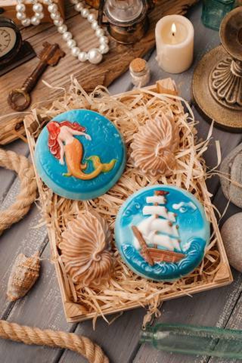 Op de afbeelding: Drie decoratieve zeepjes in een houten doos met stro. E&eacute;n zeepje heeft een zeemeermin met een staart, een ander heeft een schelp en de derde heeft een zeilboot op zee.