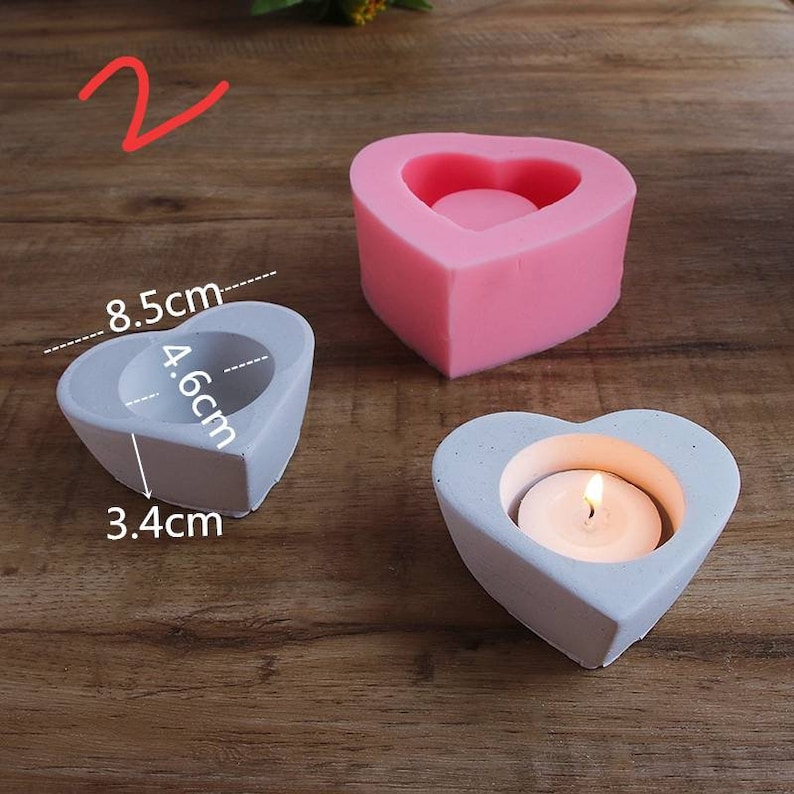 May include: Two heart-shaped candle holders, one pink and one gray, with a tealight candle in the gray holder. The pink holder is a silicone mold. The gray holder measures 8.5 cm wide, 4.6 cm tall, and 3.4 cm deep.