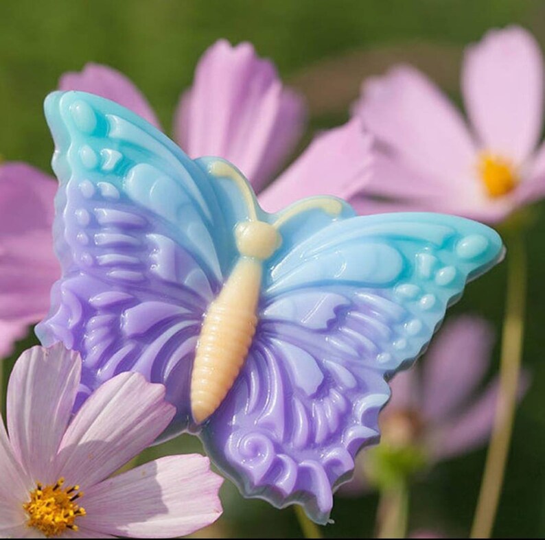 May include: A purple and blue butterfly-shaped soap with a light yellow centre. The soap is sitting on a pink flower.