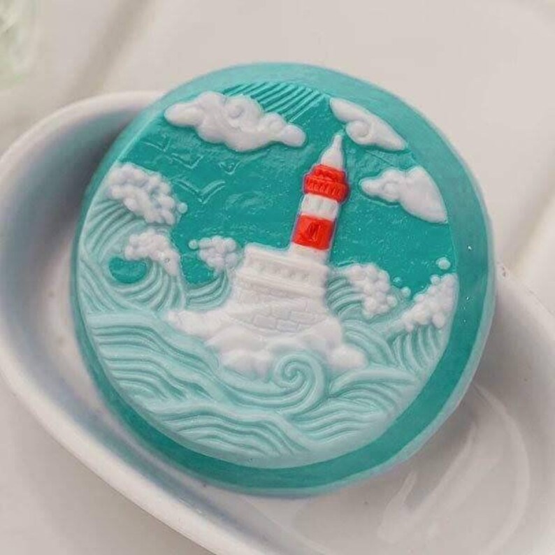 Op de afbeelding: Een ronde, turquoise zeep met een witte vuurtoren en golven die in het oppervlak zijn gegraveerd. De vuurtoren heeft een rode streep rond het midden.