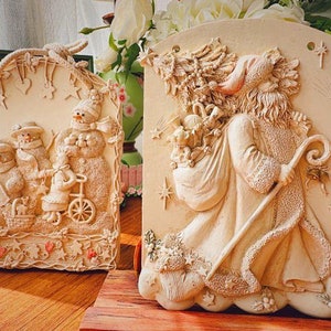 May include: Two decorative wall plaques, one with a winter scene featuring snowmen and the other with a Christmas scene featuring a figure holding a staff and a bag of gifts.