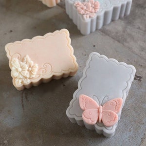 May include: Two decorative soap bars with intricate designs. One bar is light pink with a floral design and the other is grey with a butterfly design.