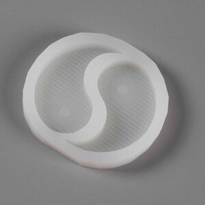 May include: White silicone mould shaped like a yin yang symbol. The mould has two separate compartments, each with a textured surface.