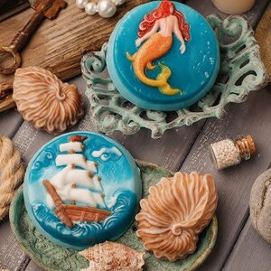 Op de afbeelding: Drie decoratieve zeepjes in de vorm van een schelp, een zeemeermin en een zeilboot. De zeepjes zijn blauw en wit met ingewikkelde details. De zeepjes worden gepresenteerd op een houten oppervlak met andere nautische decoraties.