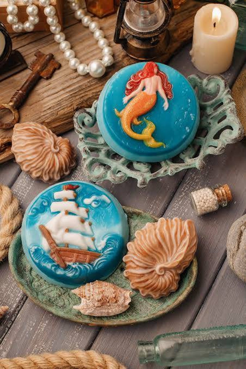Op de afbeelding: Drie decoratieve zeepjes in de vorm van een schelp, een zeemeermin en een zeilboot. De zeepjes zijn blauw en wit met ingewikkelde details. De zeepjes worden gepresenteerd op een houten oppervlak met andere nautische decoraties.