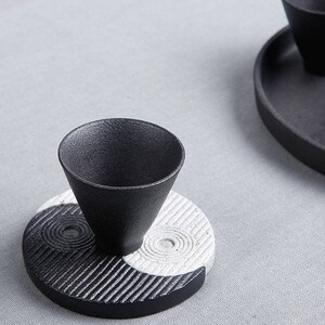 May include: Black and white ceramic teacup and saucer set with a yin yang design. The saucer is round with a black and white yin yang design. The teacup is black and sits on top of the saucer.